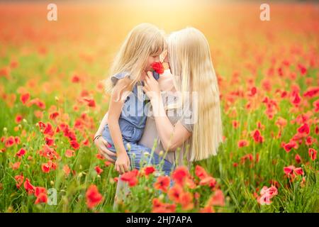 Glückliche Familie, die sich bei Sonnenuntergang auf einem schönen Mohn-Feld ausruhte. Mama hält ihre Tochter in ihren Armen inmitten einer blühenden Wiese. Familie hat Spaß. Stockfoto