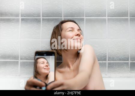 Eine Frau mittleren Alters, 40 Jahre alt, nimmt ein Bad und macht ein Selfie mit ihrem Telefon. Nette Frau, die sich im Bad erholte. Stockfoto