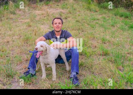 Mann mit einem weißen Hund einer Laggotto-Romagnolo-Rasse, der im Park auf Gras sitzt. Wandern mit dem Hund im Park. Hund auf der Suche nach Trüffelpilzen Stockfoto