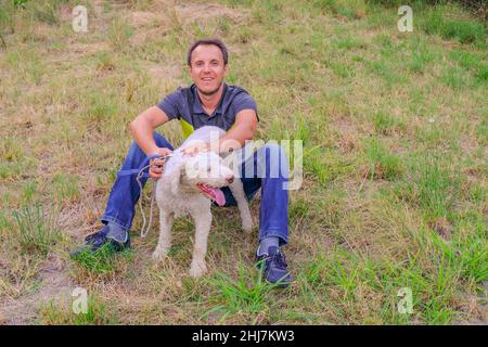 Mann mit einem weißen Hund einer Laggotto-Romagnolo-Rasse, der im Park auf Gras sitzt. Wandern mit dem Hund im Park. Hund auf der Suche nach Trüffelpilzen Stockfoto