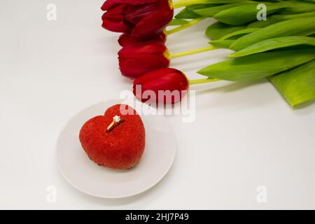 Ein Bouquet aus roten Tulpen neben einem Teller mit einem herzförmigen Kuchen, in dem ein Goldring mit einem Diamanten. Stockfoto