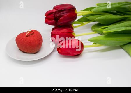 Ein Bouquet aus roten Tulpen neben einem Teller mit einem herzförmigen Kuchen, in dem ein Goldring mit einem Diamanten. Stockfoto