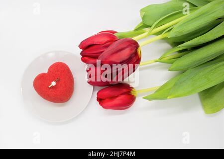 Ein Bouquet aus roten Tulpen neben einem Teller mit einem herzförmigen Kuchen, in dem ein Goldring mit einem Diamanten. Stockfoto