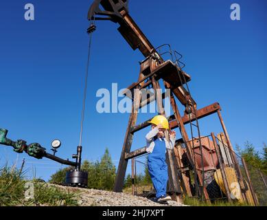 Low-Angle-Ansicht eines kleinen jungen Energietechnikers in der Nähe der Ölplattform. Boy in Overalls gegen den blauen Himmel, Petroleum-Pipeline und Pumpenheber für die Gewinnung von natürlichem Kraftstoff. Stockfoto