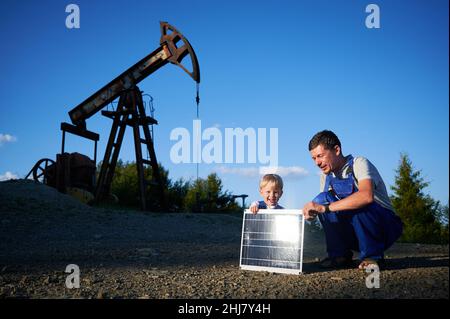 Erwachsener Mann und männliches Kind setzten sich auf Schotterplatz in der Nähe des Ölbrunnen. Sie halten Solarbatterie, Sonnenstrahlen von Solarpanel reflektiert. Zwei Varianten der Energieerzeugung mit unterschiedlichen Auswirkungen auf die Umwelt. Stockfoto