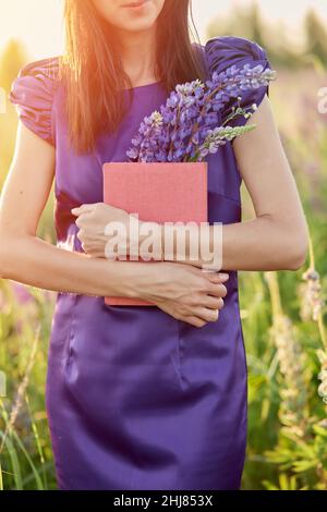 Frau im Sommerfeld mit Blumenstrauß - Lupinen, Hut und rosa Notizbuch. Nähe zur Natur, Selbsterkennungskonzept. Relax und Welll-being Konzept. Sonniger Tag, gute Laune Konzept. Stockfoto