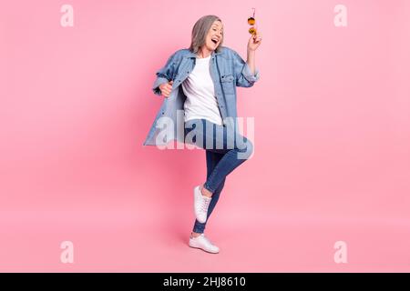 In voller Länge Foto von coolen Senior Lady Tanz tragen blaues Hemd Jeans Sneakers isoliert auf rosa Hintergrund Stockfoto