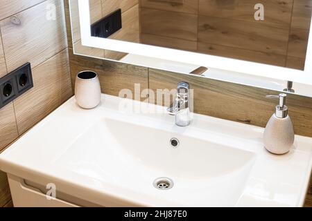 Modernes Waschbecken mit Chrom-Wasserhahn neben einem stilvollen Seifenspender. Spiegel mit integrierter LED-Beleuchtung. Stockfoto