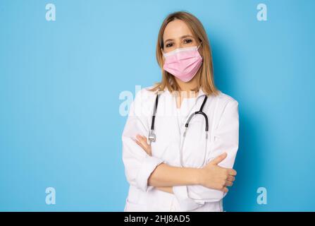 Krankenschwester stehende Arme gekreuzt. Selbstbewusste junge Frau Arzt trägt chirurgische Maske isoliert auf blauem Hintergrund. Stockfoto