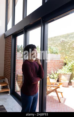 afroamerikanischer junger Mann mit der Hand am Kinn, der durch das Fenster schaut, während er zu Hause steht. Unverändert, Lebensstil, Kontemplation, Tagträumen und Kuppel Stockfoto