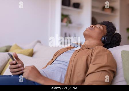 afroamerikanischer junger Mann mit geschlossenen Augen, der Musik hört, während er sich zu Hause entspannt. Unverändert, Lebensstil, Freizeitaktivitäten, drahtlose Technologie, Musik Stockfoto