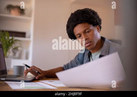 afroamerikanischer junger Geschäftsmann liest Dokument während der Arbeit mit Laptop von zu Hause aus. Unverändert, Geschäft, Daten, drahtlose Technologie und worki Stockfoto