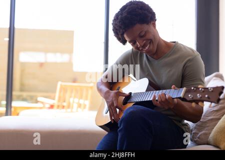 Lächelnder afroamerikanischer junger Mann, der Gitarre spielt, während er zu Hause auf dem Sofa sitzt. Unverändert, Lebensstil, Freizeitaktivitäten, Musik, Erfrischung, afro-Haare Stockfoto