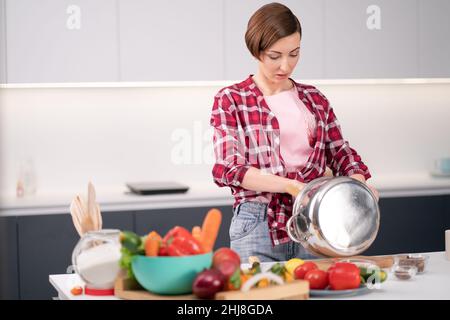 Junge Frau reinigt oder mischt Zutaten in einem Metalltopf mit Gemüse auf der Theke. Frau mit kurzen Haaren in einem roten karierten Hemd Kochen in einer modernen Küche. Home Lifestyle-Konzept. Veganes Konzept. Stockfoto