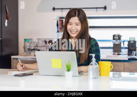 Arbeiten zu Hause in Covid-19 Pandemiekonzept, Asiatin zu Hause mit Technologie-Laptop und Handy im Küchenraum arbeiten, kurze Notiz schreiben Stockfoto