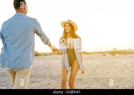 Seitenansicht eines lächelnden, glücklichen jungen Paares, zwei Familienfrauen in legerer Kleidung bei Sonnenaufgang über dem Meer, Sandstrand, Meer, Außenstrand, Sommertag, Sonnenuntergang Stockfoto