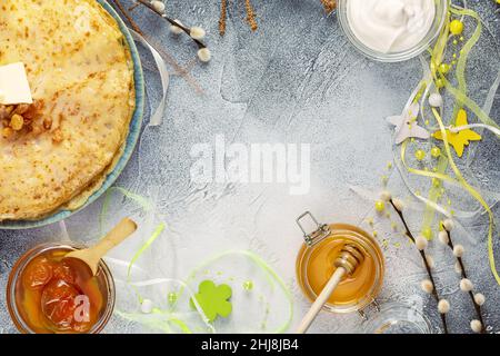 Stapel von hausgemachten frittierten Crepes mit Butter, Gläser mit Honig, Aprikosenmarmelade, Sauerrahm und Weihnachtsdekoration auf einem blauen strukturierten Hintergrund mit leeren s Stockfoto