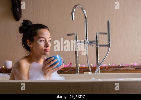 Nachdenklich biracial junge Frau schaut weg, während mit Kaffee in Seifenlauge Badewanne im Spa. Unverändert, Spa, Körperpflege, Sinnlichkeit, Lebensstil, vitiligo A Stockfoto