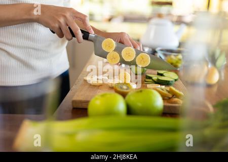 Mittelteil der biracial junge Frau hacken Bananenscheiben während der Zubereitung gesunder Saft zu Hause. Unverändert, Lebensstil und gesunde Ernährung Konzept. Stockfoto