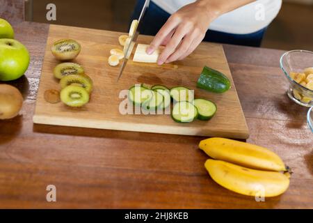 Mittelteil der biracial junge Frau hackt Banane auf Holzbrett in der Küche zu Hause. Unverändert, Lebensstil und gesunde Ernährung Konzept. Stockfoto