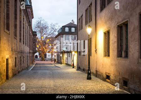 Eine leere gepflasterte Straße in der Abenddämmerung mit einem beleuchteten Baum in der Ferne, in Straßburg, Frankreich. Stockfoto