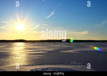 Zwei Fischer auf einem gefrorenen See fangen Fische in einem Eisloch auf dem Hintergrund des Abenduntergangs. Der Winter kommt, die Gewässer gefrieren über und machen Th Stockfoto