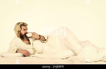 Genießen Sie den morgendlichen Kaffee. Der Körper wurde der wesentlichen Komponente für eine gute Gesundheit und Energie beraubt. Schöner Mann trinkt Kaffee. Reifer bärtiger Mann im Bett Stockfoto