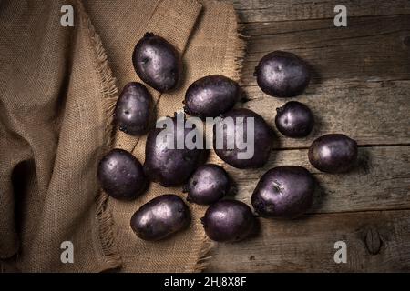 Rohe violette Vitelotte-Kartoffeln auf Holzbrettern und Quappen. Violette Kartoffeln auf Leinensacktuch. Über der Ansicht. Stockfoto