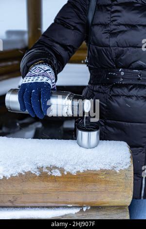 Eine Frau gießt im Winter Tee aus einer Thermoskanne auf der Straße in einen Tsakan. Hochwertige Fotos Stockfoto