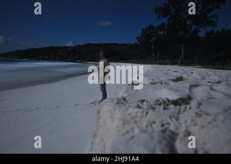 Nachtaufnahme eines Mannes an der Küste eines tropischen Strandes, beleuchtet vom Mondlicht. Langzeitbelichtung Stockfoto