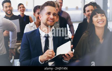 Eine kleine Aufnahme eines hübschen jungen Geschäftsmanns, der mit seinen Kollegen sitzt und ein Notizbuch in der Hand hält, während er im Büro ist Stockfoto