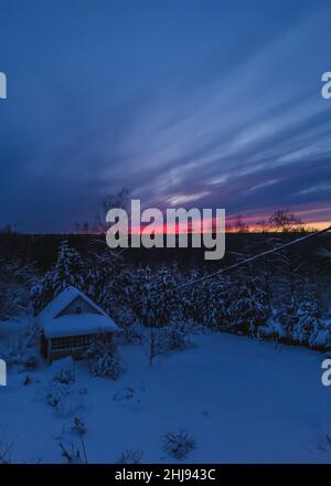 Waldhütte und Bäume im Winter Sonnenuntergang. Holzhütte im Schnee. Wald am Abendhimmel im Hintergrund. Stockfoto