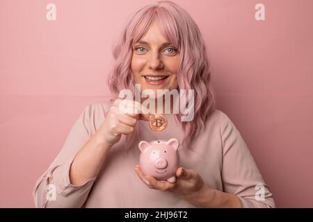 Geld sparen. Glücklich positive junge schöne attraktive kaukasische Frau Hand legt eine Bitcoin in ein Sparschwein isoliert auf rosa Hintergrund. Stockfoto