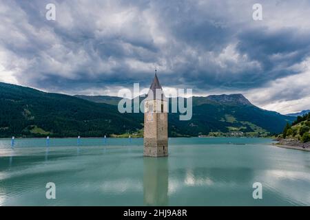 Der Glockenturm der Kirche St. Katharina ragt aus dem Wasser des Reservoirs Reschensee, bedeckt von dunklen Gewitterwolken. Stockfoto