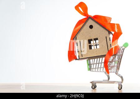 Miniatur-Push-Trolley mit einem kleinen Holzhaus mit einem roten Band Bogen isoliert auf weißem Hintergrund dekoriert. Online-Shopping für ein neues Zuhause. Stockfoto