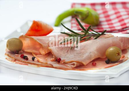 Würzige Schweineschinkenscheiben auf weißem Teller. In Scheiben geschnittener Prosciutto mit scharfer Chili, Tomaten und Oliven. Stockfoto