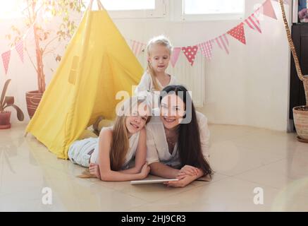 Eine glückliche Mutter mit zwei kleinen Töchtern hat Spaß, in einem gelben Wigwam im Wohnzimmer zu liegen. Stockfoto