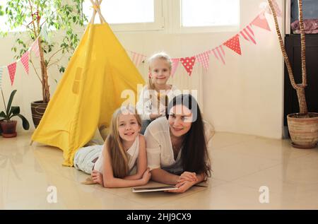 Eine glückliche Mutter mit zwei kleinen Töchtern hat Spaß, in einem gelben Wigwam im Wohnzimmer zu liegen. Stockfoto