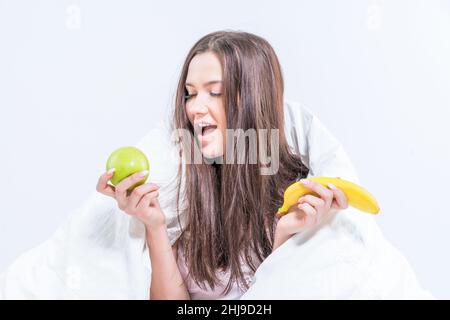 Das Mädchen sitzt auf dem Bett, in eine Decke gehüllt, hält eine Banane und schaut auf einen Apfel. Die Brünette frühstückte mit Obst. Stockfoto