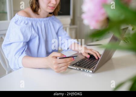 Die junge Frau in stilvoller Bluse kauft online über den Laptop und gibt Bankkartendetails hinter Blumen im Vordergrund zu Hause aus der Nähe ein Stockfoto