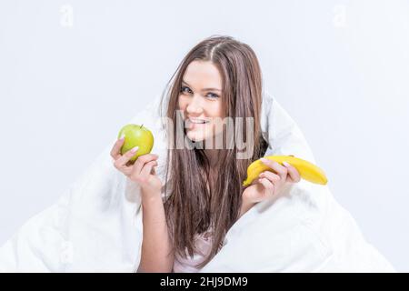 Die Brünette frühstückte mit Obst. Das Mädchen sitzt auf dem Bett, umhüllt von einer Decke und hält eine Banane und einen Apfel. Stockfoto