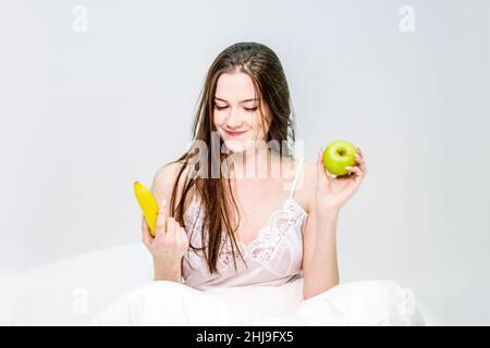 Das Mädchen sitzt auf dem Bett, umhüllt von einer Decke und hält eine Banane und einen Apfel. Die Brünette frühstückte mit Obst. Stockfoto