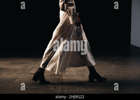 Kurze weibliche Figur in einem stilvollen langen beigefarbenen Outfit, die auf dem Laufsteg läuft und eine schwarze Handtasche hält. Details zur Fashion Week Stockfoto