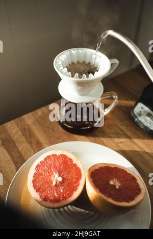 Eine Grapefruit, ein Ei und eine Banane bilden ein Frühstückslächeln auf einem weißen gesprenkelten Teller, umgeben von Utensilien, Kaffee in einem chemex und anderen Früchten Stockfoto