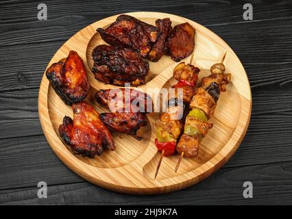 Nahaufnahme einer gemischten Grillplatte mit Hähnchenbüffelflügeln, Rindfleisch-Spareribs und Spießfleisch auf Holzbrett über schwarzem Tisch pl Stockfoto