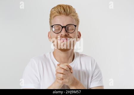 Betender junger Mann isoliert auf weißem Hintergrund. Speicherplatz kopieren. Machen Sie ein Wunsch- und Glückskonzept. Rothaariger Kerl mit rotem Bart und Brille in weißem T-Shirt Stockfoto