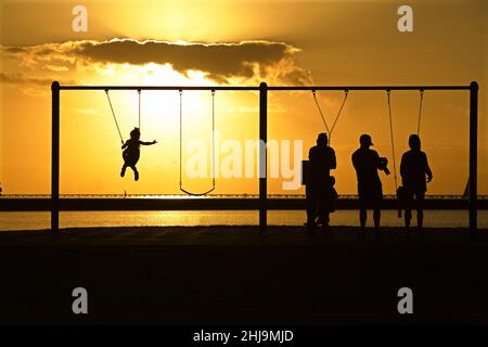 Eltern und Kinder in Silhouette spielen auf Schaukeln bei Sonnenuntergang Stockfoto
