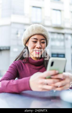 Entzückte chinesische Frau, die in der Nähe des Gebäudes in der modernen Stadt am Tisch auf der Terrasse des Cafés im Freien saß und ein modernes Smartphone browsen Stockfoto