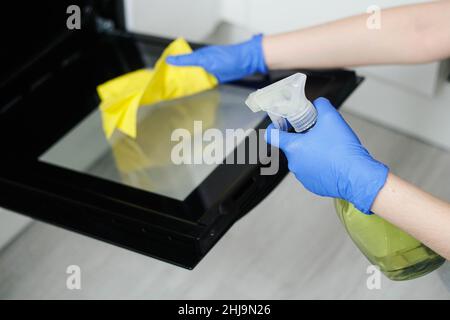 Nahaufnahme der Hände der Frau in Gummischutzhandschuhen, die den Backofen mit einem Lappen reinigen. Hausfrau spritzt Waschmittel auf die schmutzige Oberfläche des Küchenofens. Stockfoto