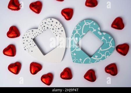 Zwei leere Bilderrahmen in Form von Herzen in Holzfarben und viele glänzende Süßigkeiten in Form von Herzen, die in Folie auf weißem Hintergrund eingewickelt sind. Bis Stockfoto